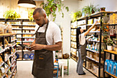 Side view of African American male seller in apron standing near shelves with groceries and looking at screen of tablet while checking information
