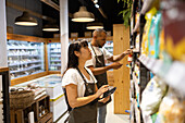 Side view of African American male and female sales staff in apron standing looking up near shelves with assorted goods and checking with help of tablet while working in shopping store