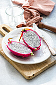 A vividly pink-skinned dragonfruit cut in half, revealing its speckled interior, presented on a white plate with a rustic wooden board beneath.