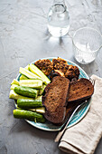 High angle of fresh organic cucumber, slices of rye sour dough bread and meat cutlets served on plate near jar of water, glass, fork and napkin against gray background