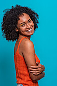 Side view of friendly African American female fashion model in casual clothes with curly hair smiling with crossed arms while standing against blue background