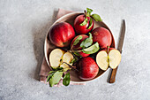 Top view of bunch of ripe red apples in plate placed near knife and cloth on gray surface against blurred background