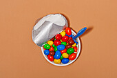 Top view of opened yoghurt container filled with colored candies and spoon against orange background