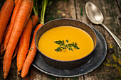 A bowl of creamy carrot soup garnished with parsley alongside raw carrots and a vintage spoon on a rustic wooden table.