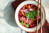 Top view of bowl with beetroot soup with onion, coriander and noodles in Asian style served on bowl and chopsticks on napkin against gray surface in daylight