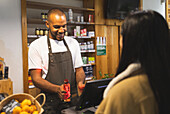 Positive young African American male seller in apron smiling and giving bottle of oil to anonymous customer while standing at counter during work
