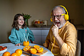 Niedliche Enkelin schaut auf einen fröhlichen Großvater in Freizeitkleidung, der Kopfhörer trägt und ein Lied über eine Buchse mit Orangen und Saft auf dem Tisch zu Hause singt