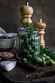 Ingredients for preparing cucamelon fermentation placed in jar placed on wooden tray near napkin and salt and pepper shakers against dark background