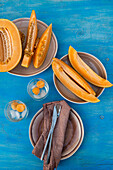 Draufsicht auf in Scheiben geschnittene frische Bio-Orangen-Moschus-Melone und Melonenkugeln in transparenten Gläsern mit Eiswürfeln, serviert auf einem Tisch mit Serviette, Gabel und Messer bei Tageslicht