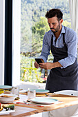 Focused Male cook in apron taking photo on smartphone of served table for breakfast in modern dining room during work