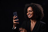 Positive Hispanic young female in outfit smiling and browsing mobile phone while standing on black background