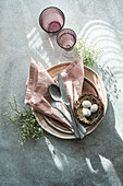 Draufsicht auf einen elegant und einfach gedeckten Ostertisch mit einem Vogelnest mit gesprenkelten Eiern, umgeben von zartem Porzellan und zartrosa Leinen