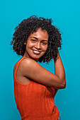 Side view of friendly African American female fashion model in casual clothes touching curly hair and smiling while standing against blue background