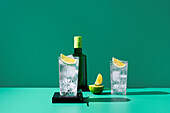 An elegant display of gin tonic in two glasses with ice and lime, alongside a bottle of gin and a halved lime, against a green backdrop.