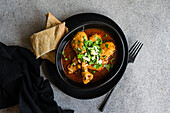 Top view of Georgian dish Chakhokhbili is stewed chicken meat in tomato sauce with spices and herbs served on black plate with some bread on gray table near fork and napkin against gray background