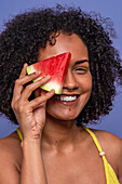Friendly charismatic African American female model smiling and looking at camera while hiding eye behind slice of sweet watermelon