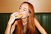 Young woman with red hair and piercing with closed eyes sitting at table and enjoy eating hamburger in restaurant