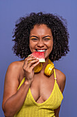 Optimistic African American female model with headphones smiling and looking at camera while biting juicy delicious watermelon in summer