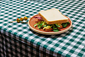Appetizing vegetarian sandwich with fresh lettuce on plate served with olives on skewer on table with checkered tablecloth