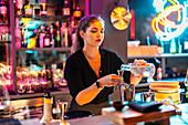 Confident beautiful young barkeeper pouring alcohol in jigger from glass bottle at bar counter while working in illuminated nightclub