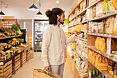 Positive side view young curly hair male customer in casual clothes standing near shelves with goods while shopping with bag in grocery shop