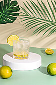 Sparkling transparent crystal glass with ice and slices of lime in cocktail drink on coaster on green surface table with limes near leaves