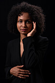 Portrait of young Hispanic female with curly hair in jacket touching face and looking at camera against black background