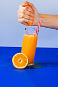 Crop anonymous person squeezing fresh refreshing orange juice into cocktail glass cup with fresh slice of orange on blue background