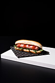 Appetizing bun with sausage and ketchup with rocket salad served table mat over white wooden board against black background in studio