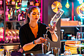 Confident beautiful young barkeeper pouring alcohol in jigger from glass bottle at bar counter while working in illuminated nightclub