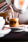 Selektiver Fokus von Milch, die in eine Glastasse mit frischem aromatischem Espresso fließt, die auf einer Untertasse steht, während ein Milchkaffee zubereitet wird
