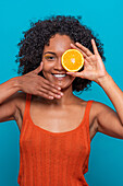 Happy young Hispanic female with curly hair covering eye with slice of orange and touching face while looking at camera against blue background