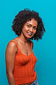 Side view of friendly African American female fashion model in casual clothes with curly hair smiling while standing against blue background