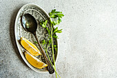 Top view of spoon with virgin olive oil, lemon slices and green parsley herb on plate against gray background