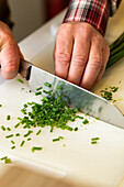 High angle of crop anonymous person's hands skillfully chopping fresh green chives on a white cutting board, with the finely cut pieces scattered across the surface