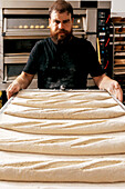 Männlicher Bäcker mit rotem Bart und schwarzem T-Shirt bereitet sich darauf vor, ein Blech mit rohem Teig in den Ofen zu schieben, um Brote in der Küche einer Bäckerei zu backen