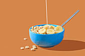 A blue bowl of cereal with banana slices being served with a splash of milk on an orange background.