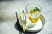 Hochformatiger Sommercocktail mit Zitronenwodka, Zitronenscheiben und wilden Minzblättern mit Eis auf einem Teller mit Strohhalmen auf einem grauen Tisch