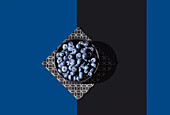Blick von oben auf eine Schale mit saftigen Blaubeeren auf einer geometrisch gemusterten Serviette vor einem kontrastierenden blauen und schwarzen diagonalen Hintergrund