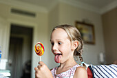 A cheerful young girl with braided hair delightedly looks at a large, colorful lollipop in a bright home setting.