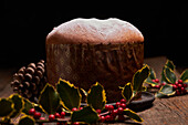 Ein wunderschön präsentierter traditioneller Panettone mit einer leichten Schicht Puderzucker, vor einem Hintergrund mit festlicher Dekoration, darunter Stechpalmenbeeren und Tannenzapfen