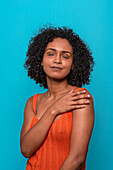 Smiling African American female model in casual clothes with dark curly hair touching shoulder with closed eyes against blue background