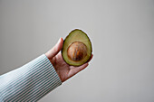 Crop anonymous person with delicious freshly sliced half of ripe avocado pear with seed held against gray background