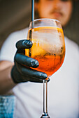 Unrecognizable bartender with gloves holding an orange cocktail with ice and a straw standing at bar in blurred background