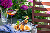 Blick von oben auf appetitliche süße Waffeln mit Pfirsichmarmelade auf rundem Tisch neben Glas mit Getränk im Garten mit blühenden lila Hortensienblüten