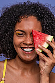Human face of friendly charismatic African American female model smiling and looking at camera while hiding eye behind slice of sweet watermelon
