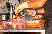 Nahaufnahme der Hände eines anonymen Kochs mit gelben Handschuhen, der gekonnt einen Oktopus auf einem Metalltisch in Scheiben schneidet