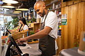 Side view positive young African American male seller in apron smiling and entering information in computer while standing at counter during work