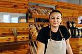 Porträt einer selbstbewussten, positiv gestimmten Verkäuferin in Schürze, die mit den Händen auf dem Tresen steht, während frisch gebackene Brote auf Holzregalen in einer Bäckerei angeordnet sind