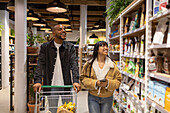 Cheerful young multiracial couple in casual wear smiling and carrying shopping trolley while choosing goods from shelves in supermarket together
